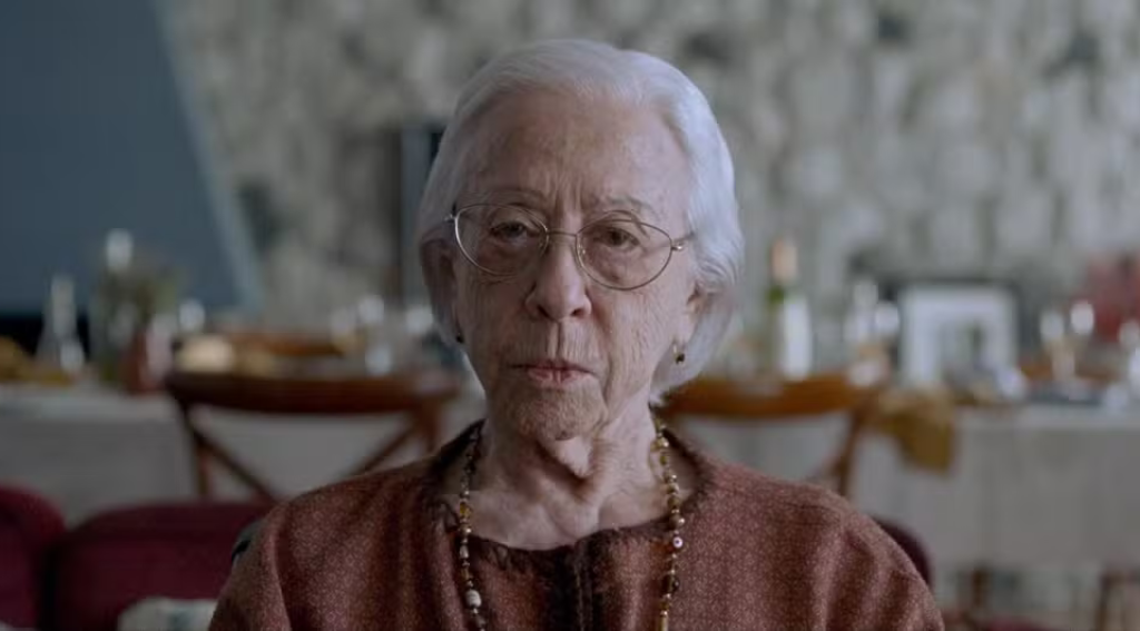 Cena do filme Ainda Estou Aqui Na imagem, Eunice Paiva, mulher branca na faixa dos 95 anos, de cabelo curto branco, está olhando para frente com expressão de desorientação. Ela veste uma roupa marrom e usa um colar de bolinhas. Usa óculos redondos. Ao fundo há uma mesa, desafogada, com garrafas, copos e pratos. 
