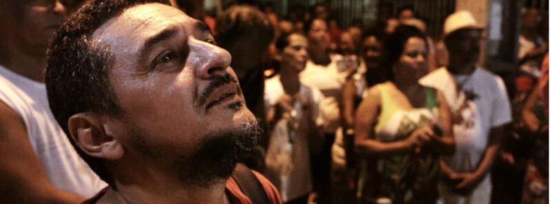 Cena de O Palhaço de cara limpa. Na imagem, um homem pardo aparece olhando para o céu. Ele veste camiseta vermelha e está no meio de uma manifestação que acontece a noite entre diversas pessoas. Sua feição é triste e tem lágrimas no canto do olho. 