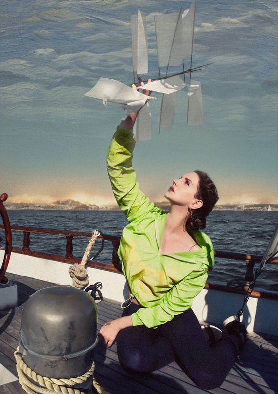 Lana Del Rey está ajoelhada no convés de um barco, usando uma blusa verde neon. Ela olha para cima com uma expressão contemplativa enquanto segura um pequeno modelo de barco de papel com várias velas, levantando-o em direção ao céu. O fundo da imagem mostra o mar tranquilo e um céu parcialmente nublado. A luz suave e os tons de azul e verde criam uma atmosfera de sonho e serenidade.