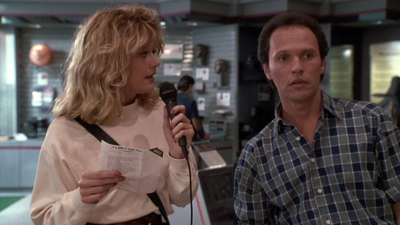 Cena do filme Harry e Sally , com duas pessoas em uma loja de eletrodomésticos. A esquerda temos Sally, uma mulher com cabelo loiro e ondulado, ela está segurando um microfone e um papel na mão,parecia estar cantando ou  falando. Ela veste uma blusa branca de manga comprida e tem uma bolsa preta à tiracolo. Ao lado dela temos Harry,  um homem de cabelo curto que está com uma expressão de surpresa ou confusa, ele veste uma camisa de flanela xadrez em tons de azul. O fundo é de uma loja de eletrodomésticos, com pessoas ao fundo e itens expostos