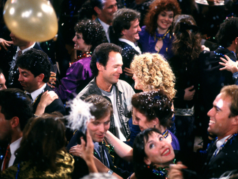 Em uma cena do filme “Harry e Sally” há uma multidão de pessoas em uma festa, onde confetes e balões enfeitam o ambiente. No centro da imagem, há Harry homem e Sally uma mulher e estão dançando juntos, sorrindo um para o outro. Harry tem cabelo curto e usa uma jaqueta clara, enquanto Sally tem cabelos loiros cacheados e veste uma roupa elegante. Ao redor deles, outros casais dançam e se abraçam, em clima de celebração e alegria. A cena é cheia de cores vibrantes e expressões felizes