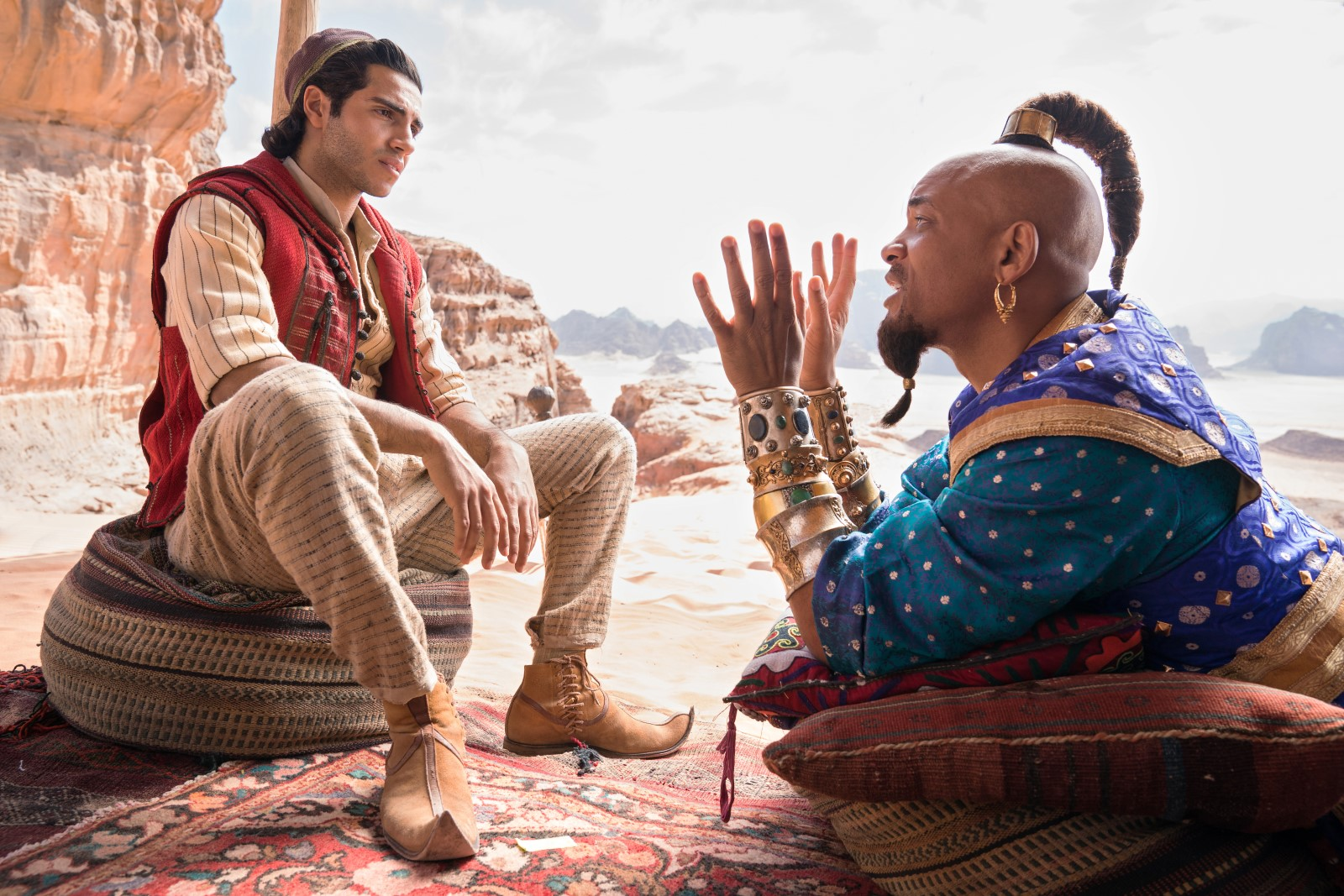 cena do live-action Aladdin. Aladdin e o Gênio estão no deserto conversando. Aladdin está à direita, de frente para o Gênio que está à esquerda; o primeiro está sentado em um assento de pano (um puff) e mantém os antebraços apoiados nas coxas com as mãos para baixo, entre as pernas. Ele olha para o Gênio com um semblante concentrado, levemente confuso. O Gênio está deitado, de bruços, em um puff com duas almofadas em cima. Ele se apoia nos cotovelos e levanta as mãos abertas na altura do rosto, gesticulando para Aladdin. Eles estão em cima de um tapete, Aladdin veste uma calça bege com listras pretas, uma camisa da mesma cor e estampa com uma sobreposição semelhante a um colete na cor vermelha. O gênio utiliza uma túnica azul e tem braceletes de ouro com pedras incrustadas, que no filme funcionam como as algemas que o ligam à lâmpada mágica.