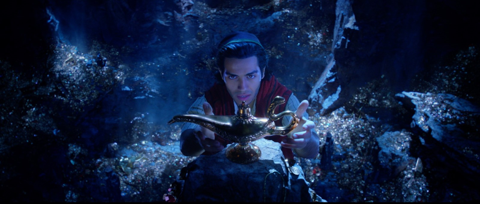 Cena do live-action Aladdin. Mena Massoud, intérprete de Aladdin, está centralizado e estende suas mãos para pegar a lâmpada mágica. O enquadramento é feito de cima para baixo, a lâmpada se destaca, um pouco mais alta do que o personagem, porém em desfoco. Aladdin olha fixamente para ela e abre suavemente sua boca, quase como em um começo de um sorriso. Ele está dentro de uma caverna cheia de ouro e tesouros; o ambiente possui um tom de azul escuro e uma luz ilumina o persongem e parte do objeto pelo lado direito. Os tesouros reluzem e criam um contraste no ambiente escuro.