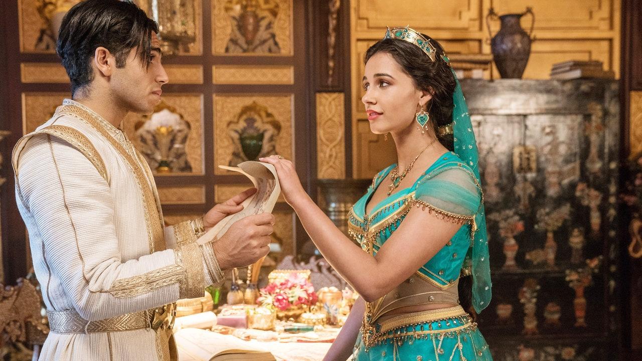 cena do live-action Aladdin. A princesa Jasmine, interpretada por Naomi Scott, e Aladdin, interpretado por Mena Massoud, estão em pé, um de frente para o outro e de perfil para a câmera. Aladdin está disfarçado de Príncipe Ali, com vestes brancas, e segura um pergaminho aberto com ambas as mãos. Jasmine, à direita, veste seu vestido característico de seda turquesa e joias que combinam com a roupa, um colar e brinco de pedras (ambos de tamanho grande) e uma coroa dourada com também pedras turquesas incrustadas. Ela abaixa, delicadamente com uma das mãos, o pergaminho que o rapaz segura. A princesa mantém um semblante calmo, enquanto Aladdin levanta as sobrancelhas e morde o lábio inferior. Eles estão em uma das salas do palácio, atrás dos jovens há uma mesa com objetos de ouro e livros. A parede é ornamentada, também de ouro e com espaços que são ocupados por o que aparenta ser relíquias. O plano atrás de Aladdin e Jasmine está em desfoque.