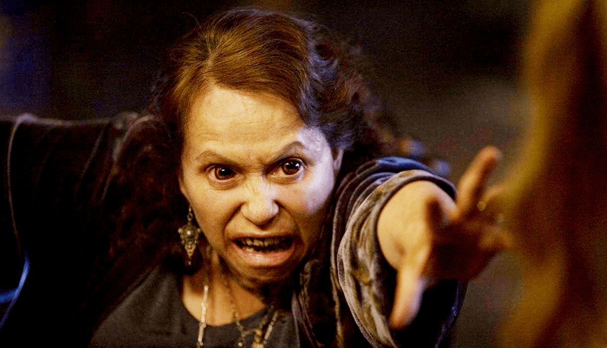 Cena do filme Arraste-Me para o InfernoNa cena, a personagem Shaun San Dena, interpretada por Adriana Barraza, está com o braço esticado para frente e a mão aberta, pronto para agarrar a personagem Chris Brown, que está de frente para Shaun, desfocada pela câmera no canto direito. Shaun está possuída, por isso seus olhos estão dilatados e os dentes afiados. Ela está com a expressão no rosto de raiva. Shaun veste roupas largas de cor marrom, possui cabelos longos na cor castanha. 
