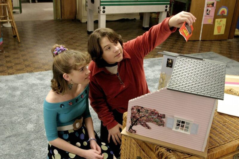 Cena do filme De Repente trinta. Na imagem estão os atores Christa B. Allen e Sean Marquette, sentados no chão sob um carpete cinza,em volta de uma casa de bonecas cor de rosa. A atriz interpreta Jenna Rink, ela é uma garota branca, com cabelos loiros presos e uma franja volumosa. Ela veste um top azul cravejado com pedras redondas, um cinto com o fecho em formato de flor e uma saia preta com bolinhas amarelas, roxas e azuis. Do seu lado esquerdo, está o ator mirim, um garoto branco com cabelos castanhos lisos, ele veste um casaco vermelho de ziper. Ela está despejando a purpurina de um saquinho laranja, sobre a casa de bonecas. 