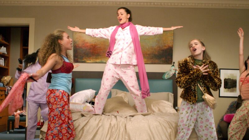 Cena do filme De repente 30. A atriz Garner interpretando Jenna está em um quarto, em pé sobre a cama. Ela está com os braços e lábios abertos, como se cantasse uma música. Ela veste um conjunto de pijama cor de rosa e um cachecol rosa. Seus cabelos castanhos estão presos em um rabo de cavalo. Em torno da cama vemos quatros garotas com pijamas e roupas engraçadas, como casaco de pele animal print, sutiã por cima da camiseta e bolsas com pijama. o quarto tem as parede bege e um quadro horizontal acima da cama. 