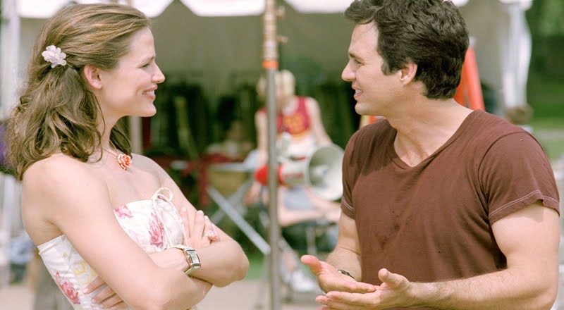 Cena do filme De Repente 30. Um close dos atores Jennifer Garner e Mark Ruffalo conversando. Do lado esquerdo a atriz Garner interpreta Jenna Rink, uma mulher branca de cabelos castanhos, presos na lateral por uma presilha em forma de flor branca. Ela veste um vestido tomara que caia branco florido rosa, e usa um colar em formato de flor pequeno e laranja. Seus prazos estão cruzados sobre o peito e em um dos seus pulso ela veste um relógio. Ela está sorrindo, olhando para o ator Ruffalo, que interpreta Matt Flamhaff. Ele é um homem branco com cabelos castanhos escuros, veste uma camiseta marrom e está gesticulando com as mãos. ao fundo se-se uma tenta com araras e cabides com roupas, uma pessoa é vista em desfoque, próximo a equipamentos de filmagem como um autofalante. 