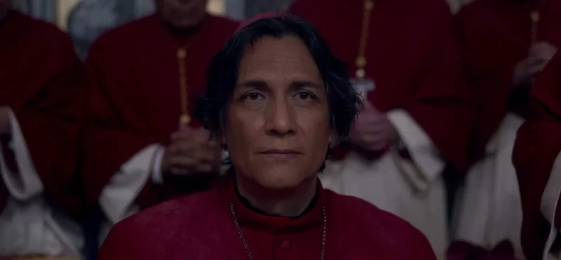 Texto Alt: Cena do filme Conclave Na imagem, o personagem Cardeal Benítez está olhando para frente com uma expressão séria. Ele veste roupas de cardeal da igreja católica, uma batina vermelha e branca; em seu pescoço há um terço fino prateado. Atrás dele, há outros cardeais, eles estão em pé, com as mãos juntas em posição de oração. Também vestem batinas e usam terços. Benítez é um homem na faixa dos 50 anos, de pele escura, mexicano, e com cabelos escuros. 