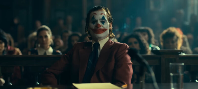 Joaquin Phoenix está vestido como o Coringa. Ele está sentado em frente a uma mesa dentro de um tribunal, atrás dele está o público que assiste a audiência.