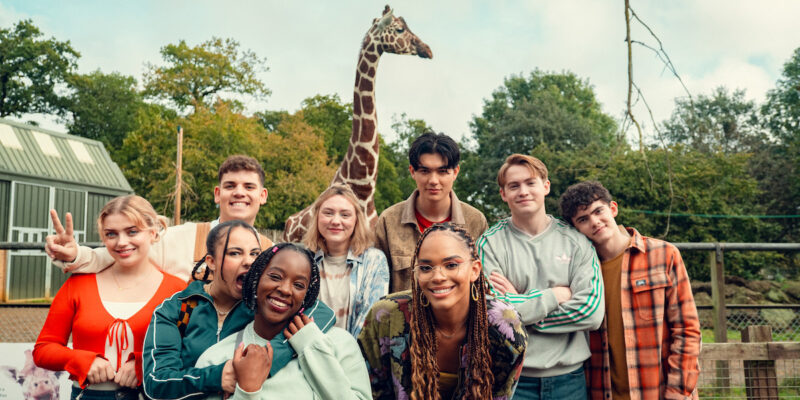 A imagem mostra um grupo de nove adolescentes posando juntos para uma foto em um ambiente ao ar livre, com uma girafa ao fundo. Eles estão sorrindo e parecem felizes, em um zoológico com árvores e construções ao fundo. O grupo é diverso, com diferentes estilos e roupas coloridas.