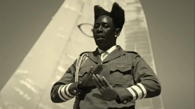 Na imagem, Tyler, The Creator veste um uniforme de soldado completo com acessórios que complementam o traje militar. Seu cabelo está dividido em duas partes, e ele aparece gesticulando enquanto performa uma música. O cenário é um avião antigo do exército dos Estados Unidos, onde Tyler está posicionado sobre uma das asas, criando uma atmosfera de intensidade e nostalgia histórica.