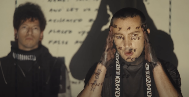 Cena do clipe Overcompensate, da banda Twenty One Pilots. Nele vemos, na parte direita da imagem, Tyler Joseph, um homem branco de cabelos raspados. Tyler veste uma camiseta preta, uma faixa escrito Clancy sobre seu pescoço e suas mãos estão pintadas de preto. Já Josh, um homem branc de cabelos pretos, veste uma camiseta preta de gola alto, com detalhe no pescoço escrito Torchebreaker. Eles estão em uma sala de fundo branco, com um slide sobre eles, no qual tem grafias escrtias a mão.