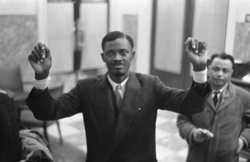 Cena do filme Trilha Sonora para um Golpe de EstadoNo centro da imagem, está Patrice Lumumba, ele está com as duas mãos erguidas mostrando o pulso enfaixado. Ele está com uma expressão séria, veste terno e gravata. Lumumba é um homem adulto de pele negra, seu cabelo é curto e ele tem bigode e cavanhaque. No lado direito, há um homem, também usando terno. 