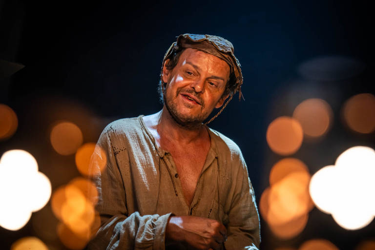 Fotografia de João Grilo iluminado pelas velas da igreja da cidade, em sua roupa velha e suja e seu chapéu da cultura nordestina enquanto performa suas malandragens.