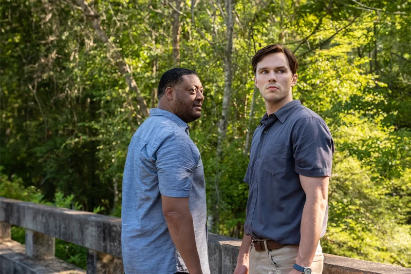 O cenário é de uma ponte, com árvores no fundo. Na imagem estão Cedric Yarbrough e Nicholas Hoult. Cedric está com uma camisa azul clara, ele está de lado e olha fixamente para Nicholas Hoult. Nicholas Hoult usa uma blusa azul, ele está de lado e evita o olhar de Cedric.