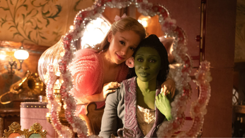 Cena do filme Wicked. As personagens Glinda e Elphaba se olham em um espelho. Glinda, branca, loira e vestida de rosa, apoia as mãos sobre os ombros de Elphaba, de pele verde, cabelos pretos em tranças finas e usando um casaco cinza e roxo sobre uma blusa branca