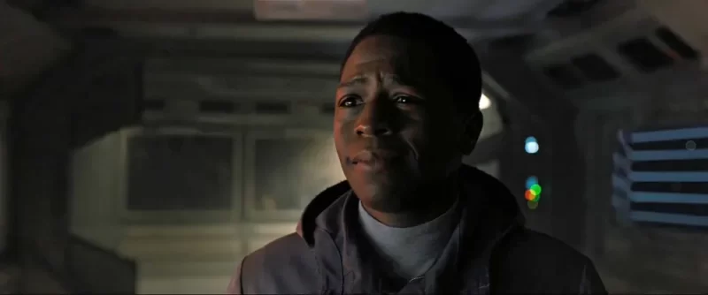 Cena do filme Alien: RomulusNa imagem, o personagem Andy está centralizado, sua expressão é de aflição. Ele é um homem adulto na faixa dos 30 anos, de pele negra e cabelos crespos curtos. Ele está vestindo uma jaqueta cinza com gola e por baixo uma roupa branca. O cenário é de uma nave, ela é acinzentada e há pouca iluminação. 
