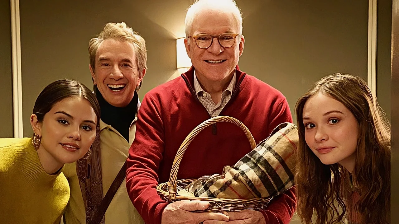 Cena da terceira temporada de Only Murders in the Building. Na imagem vemos Mabel, Oliver, Charles e Lucy encarando algo através do batente da porta. Mabel, mulher branca de cabelos castanhos escuros, veste um suéter de lã na cor mostarda. Oliver, homem branco com cabelos claros e grisalhos, usa camiseta de gola alta sobreposta por uma camisa bege. Charles, homem branco de cabelos brancos, veste uma camisa xadrez vermelha sobreposta por um suéter também na cor vermelha. Lucy, garota branca com cabelos castanhos claros, usa uma blusa vermelha. Em suas mãos, Charles segura uma cesta com uma coberta com tecido de caxemira. Ao fundo a parede está na cor marrom, uma luminária encontra-se posicionada ao centro.