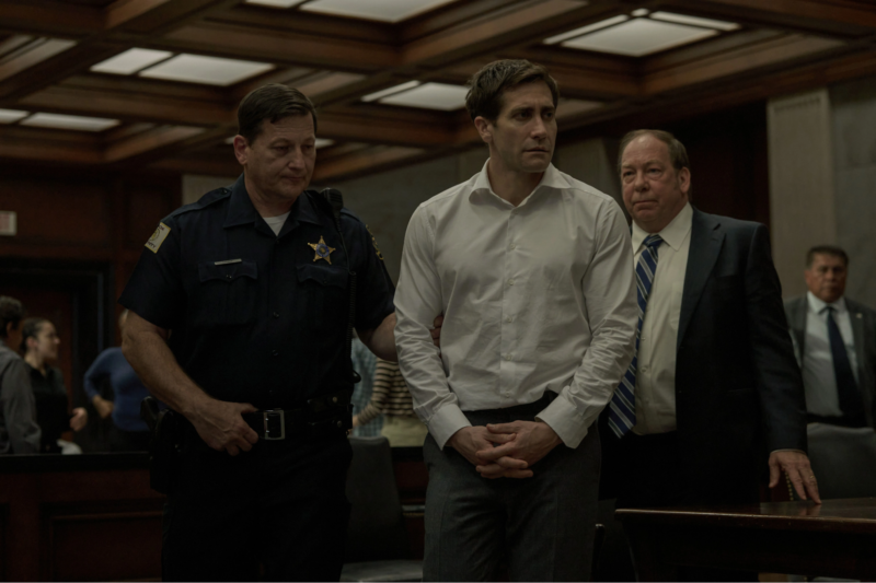 A imagem mostra uma cena em um tribunal. Um homem, vestido com uma camisa branca e calças cinzas, está sendo conduzido por um policial, que usa um uniforme azul escuro com um distintivo. O homem parece preocupado e tem as mãos juntas na frente do corpo. Atrás deles, um homem de terno escuro com uma gravata listrada observa. O ambiente é bem iluminado, com um teto de painéis de madeira e várias pessoas visíveis ao fundo.