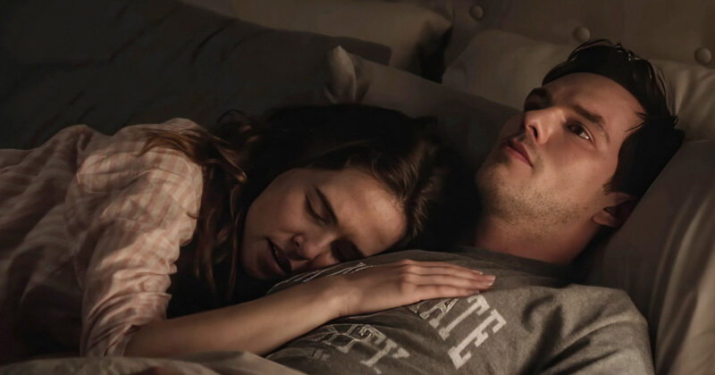 Na cena estão Nicholas Hoult e Zoey Deutch. Ambos estão na cama. Ela está deitada com a cabeça e a mão no peito dele. Ele olha fixamente para o teto.