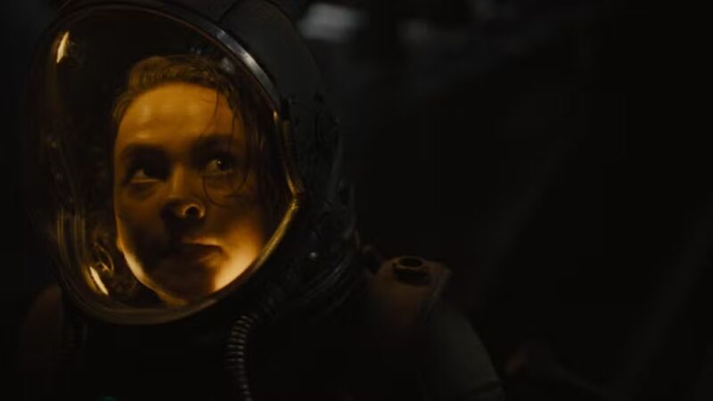 Cena do filme Alien: RomulusNa imagem, a personagem Rain está no canto esquerdo, vestindo um uniforme de astronauta e o capacete. O cenário é todo escuro e dentro do capacete há uma luz amarelada iluminando o rosto de Rain. A expressão dela é atenta ao perigo. Rain é uma mulher adulta na faixa dos 25 anos, de pele clara e cabelos escuros.
