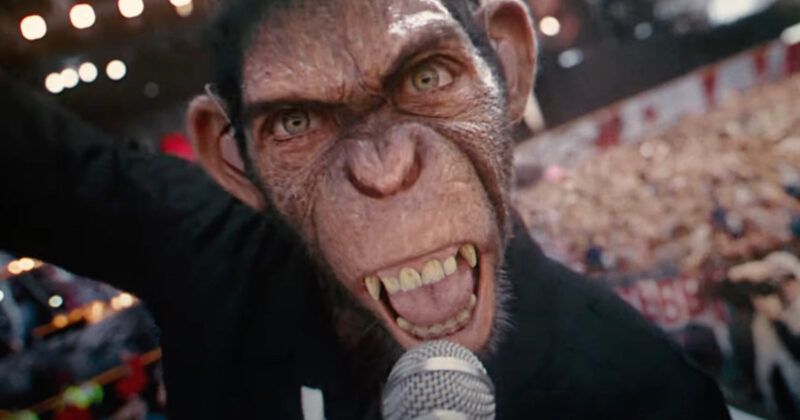 Cena do filme Better Man - A História de Robbie WilliamsNa imagem, o personagem Robbie está cantando de forma feroz para a câmera. Robbie é um chimpanzé, com olhos verdes, pelos pretos e dentes pontudos e amarelados. Ele veste uma roupa de mangas longas na cor preta. Ele canta segurando um microfone. O cenário é de um show, em desfoque, há luzes e muitas pessoas na plateia. 