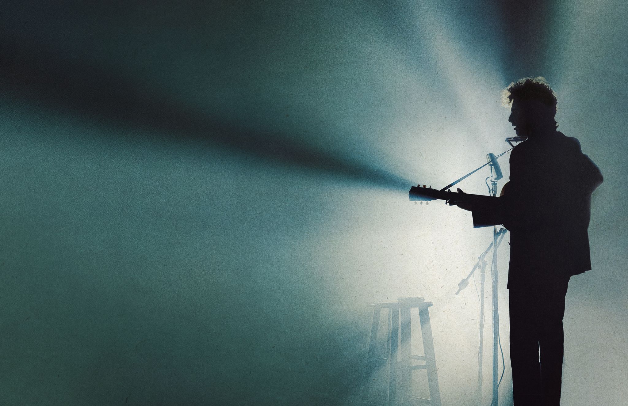 Cena do filme Um Completo Desconhecido. Na cena, há uma silhueta de um músico em um palco, iluminado por uma luz em foco, tocando guitarra. A composição da imagem é simples e focada, com o músico no centro, em destaque. O artista está posicionado em um palco, possivelmente numa apresentação. A iluminação, direcionada ao músico, cria um contraste forte entre a figura escura e o fundo levemente iluminado. As linhas do corpo do músico, bem como a guitarra e o microfone, ficam bem definidas, sem detalhes excessivos. A posição do músico sugere movimento e ação, como de uma apresentação musical.