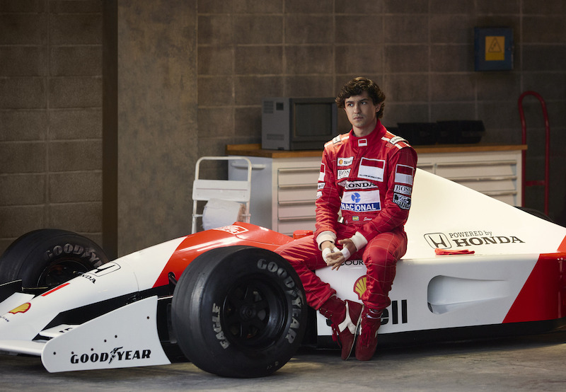 Fotografia de Gabriel Leone, homem branco de barba raspada e cabelo preto e sobrancelhas grossas interpretando Ayrton Senna, vestido no tradicional macacão vermelho com seus patrocinadores e sentado em cima de sua McLaren MP4/5 branca e vermelha com os adesivos de patrocínios tanto do piloto quanto da equipe britânica.