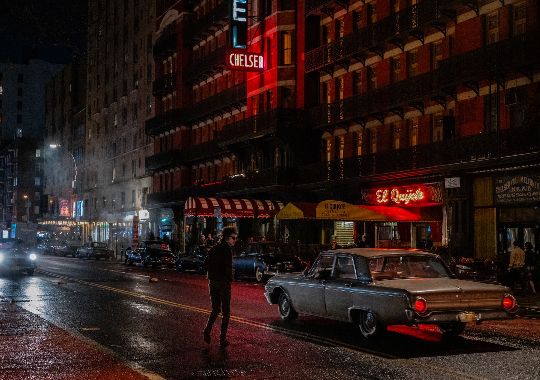 Cena do filme Um Completo Desconhecido. Na cena, uma rua noturna movimentada, com edifícios de tijolos vermelhos e letreiros luminosos em vermelho, ilustra um momento de uma cidade movimentada à noite. O foco principal é um homem que caminha na rua. Ele está vestido com roupas escuras e parece estar caminhando com uma expressão neutra. Os carros estacionados ao longo da rua são de vários modelos e estão em diferentes pontos de estacionamento.