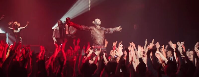 Cena do filme Better Man - A História de Robbie WilliamsNa imagem, Robbie Williams está no centro de um palco, sendo aclamado por muitas pessoas, que levantam a mão para o alto em direção ao personagem. O cenário é de um show. Robbie está com os braços abertos, rosto para cima e boca aberta. Há uma luz sob ele. A plateia está dividida por cores, o lado esquerdo iluminado por vermelho e o direito por uma luz branca. Atrás de Williams há dançarinas e outros cantores. Robbie é um chimpanzé, com olhos verdes e pelos pretos. 