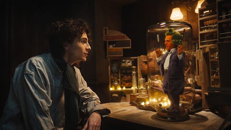 Cena do filme Wonka. Na imagem temos Willy Wonka (Timothée Chalamet), um jovem branco, de cabelo castanho ondulado. Ele veste uma camiseta listrada (branca e azul) de mangas longas e um lenço preto ao redor do pescoço. Ele está sentado enquanto conversa com um Oompa-Loompa (Hugh Grant), um ser místico laranja de porte pequeno. Ele está vestindo um conjunto roxo xadrez e um sapato de elfo na mesma tonalidade das roupas. Ambos estão em um quarto escuro, iluminado por algumas lâmpadas. No fundo é possível observar os ingredientes que o Willy Wonka utiliza para fazer os seus chocolates. 