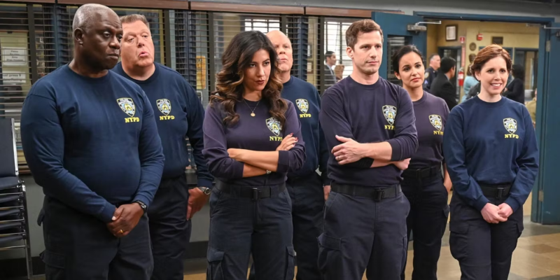 Cena da série Brooklyn Nine-Nine. Na imagem, há sete pessoas alinhadas em uma sala da delegacia, todas vestindo camisetas de manga comprida azul-marinho com o distintivo da polícia de Nova York no lado esquerdo do peito. À esquerda, Holt, que é um homem negro e careca, mantém uma expressão séria, enquanto ao seu lado Scully, um homem branco de cabelo curto grisalho, parece surpreso, com a boca entreaberta. No centro, Rosa, uma mulher de cabelos longos e escuros, está de braços cruzados, transmitindo confiança. Ao lado dela, Jake, um homem branco de cabelo castanho curto também mantém os braços cruzados, com uma expressão firme. Mais à direita, Amy, uma mulher de cabelos escuros presos sorri levemente, e ao seu lado, Hitchcock, um homem branco careca aparece parcialmente visível e parece confuso. À extrema direita, Debbie, uma mulher branca de cabelos ruivos curtos sorri abertamente, parecendo a mais descontraída do grupo. Todos estão observando algo à frente, sugerindo que aguardam alguma instrução ou estão prestes a entrar em ação.