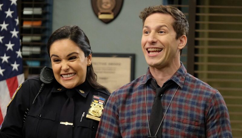 Cena da série Brooklyn Nine-Nine. Na imagem, Jake, um homem branco de cabelos castanhos curtos veste uma camisa xadrez azul e vermelha e sorri abertamente, Ao seu lado, Amy, uma mulher de cabelos escuros presos, veste um uniforme policial preto e também sorri animadamente. Ao fundo, nota-se uma bandeira dos EUA, uma parede azul clara e duas janelas.