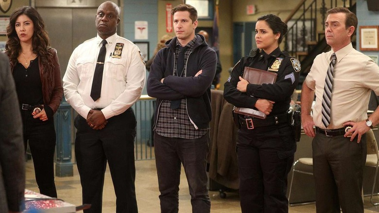 Cena da série Brooklyn Nine-Nine. Na imagem, um grupo de cinco pessoas está reunido em uma delegacia, todos com expressões sérias e posturas fechadas. À esquerda, Rosa, uma mulher de cabelos longos e escuros, veste uma jaqueta preta sobre uma blusa preta. Ao lado dela, Holt, um homem negro, careca, usa uma camisa branca de mangas longas com insígnias da polícia e uma gravata preta, transmitindo uma presença autoritária. No centro, Jake, um homem branco de cabelos curtos e castanhos veste uma camisa xadrez azul sob um moletom escuro, com os braços cruzados. À sua direita, Amy, uma mulher de cabelos escuros presos, veste um uniforme policial preto e segura uma prancheta contra o peito. Por fim, à extrema direita, Charles, um homem branco de cabelo curto veste uma camisa bege de manga curta com uma gravata marrom e mantém as mãos nos quadris, parecendo atento à conversa.