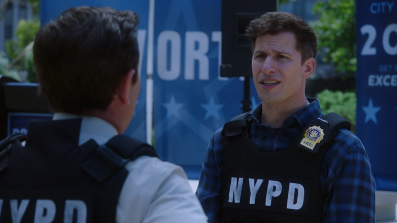 Cena da série Brooklyn Nine-Nine. Na imagem, Jake, um homem branco de cabelos castanhos curtos veste uma camisa xadrez azul e um colete à prova de balas com a sigla "NYPD" estampada na frente. Ele também tem um distintivo preso ao colete e exibe uma expressão confusa ou preocupada enquanto conversa com outra pessoa, que aparece de costas. Essa segunda pessoa também veste um colete idêntico, com a mesma sigla policial, e parece estar ouvindo atentamente o que o homem à sua frente está dizendo. O cenário ao fundo sugere um evento ou cerimônia policial, com faixas azuis e inscrições que remetem a uma campanha ou evento oficial.