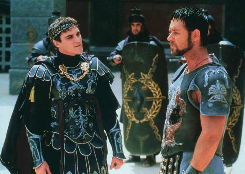 Cena do filme Gladiador. Na imagem, o imperador Cômodo, interpretado por Joaquin Phoenix, encara o gladiador Maximus dentro da arena. Cômodo veste uma armadura negra ornamentada com detalhes dourados e pratas, com uma coroa de louros, enquanto Máximus usa uma armadura prateada danificada e está ferido. Ao fundo, guardas romanos armados com escudos observam a cena. Phoenix é um homem branco, jovem e de cabelos pretos.