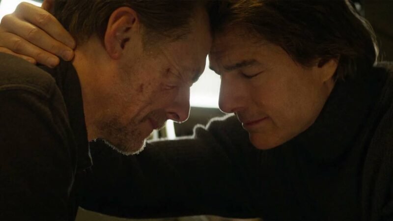 Cena do filme Missão: Impossível - O Acerto FinalNa imagem, os personagens Benji (à esquerda) e Ethan Hunt (à direita) estão com as testas encostadas, em sinal de afeto. Benji é um homem na faixa dos 50 anos, ruivo, de pele branca. Ele possui arranhões na bochecha e está com os olhos fechados, segurando o choro. Já Ethan Hunt é um homem na faixa dos 60 anos, de cabelos na altura do pescoço, cor escura e de pele clara. Ele também está com os olhos fechados. Ambos vestem roupa escura. Do cenário não dá para ver nada, apenas uma luz branca que está ao fundo.