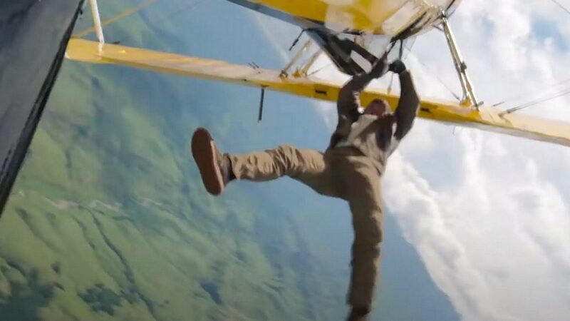 Texto Alt: Cena do filme Missão: Impossível - O Acerto FinalNa imagem, Ethan Hunt está pendurado em um avião de cor amarela. Ele está nas alturas, no horizonte é possível ver nuvens e vegetação. O personagem veste uma calça bege, camiseta branca, tênis e jaqueta marrom. Ele é um homem branco, na faixa dos 60 anos.