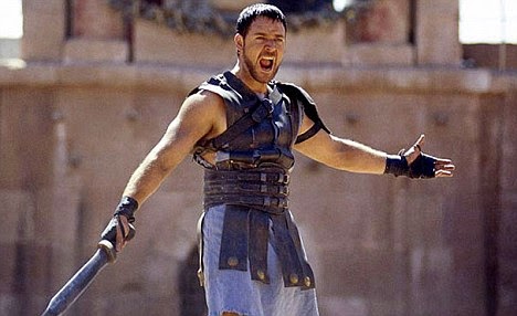 Cena do filme Gladiador. Na imagem, Maximus está em uma arena romana, sob a luz intensa do sol. Ele está ao centro da imagem, gritando com expressão de fúria. Enquanto segura uma espada na mão direita e mantém o braço esquerdo estendido em desafio. Usa uma armadura de couro escura e está cercado pela grandiosidade da arena.