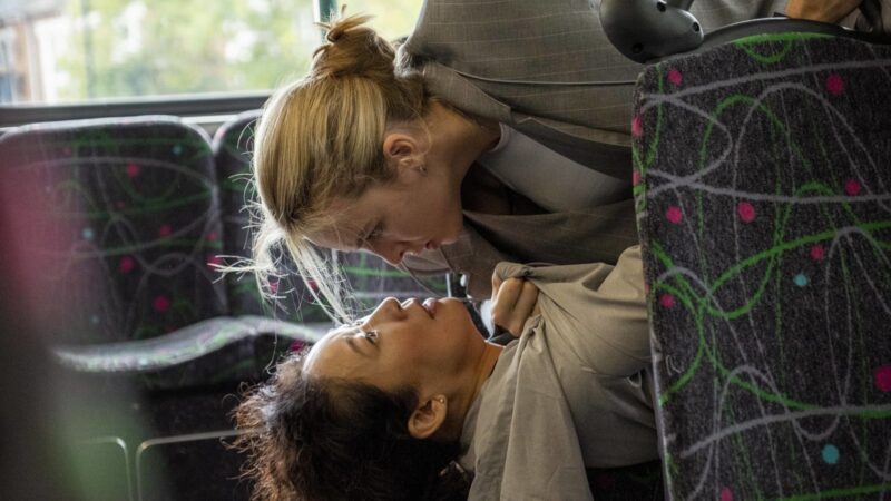 Cena de Killing Eve. Ambas personagens, Villanelle e Eve Polastri, estão dentro de um ônibus. Villanelle, mulher branca de cabelos loiros presos em um coque, veste um blazer cinza e uma camiseta branca. Ela está em cima de Eve, enquanto a segura pelo colarinho. Eve é uma mulher com ascendência sul-coreana e cabelos escuros cacheados. Ela veste um casaco cinza claro de tecido leve e está embaixo de Villanelle. Eve aparenta assustada e Villanelle a olha com ira. Os rostos de ambas estão muito próximos.