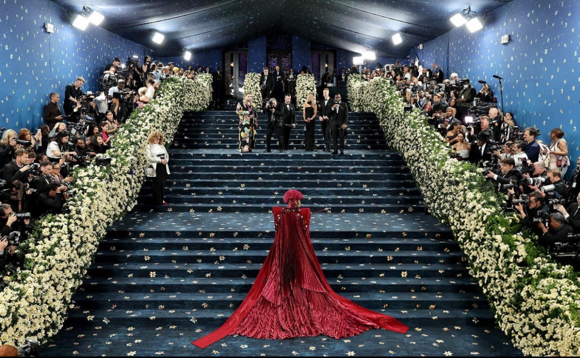 Visão ampla da icônica escadaria do Met Gala 2025, coberta por um luxuoso tapete azul profundo com detalhes dourados que lembram pequenas flores. As laterais da escada são decoradas com densos arranjos de flores brancas, formando corredores naturais que conduzem os convidados até o topo. No centro da imagem, uma figura vestida com um deslumbrante vestido vermelho de cauda longa e volumosa sobe as escadas, criando um contraste marcante com o azul do tapete. Centenas de fotógrafos e convidados observam atentamente dos dois lados da escadaria. A atmosfera é sofisticada e cinematográfica, destacando a grandiosidade do evento.