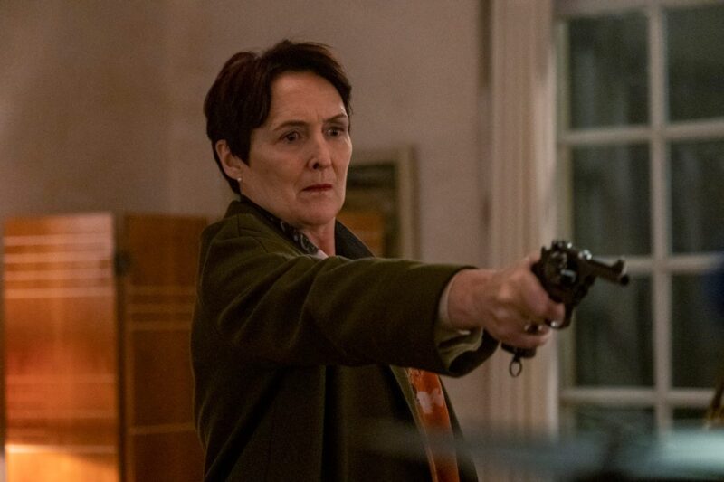 Cena de Killing Eve. Na imagem, está Carolyn Martens, personagem da série. Ela é uma mulher de meia-idade, branca, de cabelos curtos e avermelhados. Carolyn, com feição brava, aponta uma arma, enquanto veste um casaco verde-escuro e uma echarpe com fundo vermelho. Atrás dela, vemos uma parede branca com algumas marcas amareladas, as dobradiças de um suposto armário, um quadro e uma grande janela. É noite.