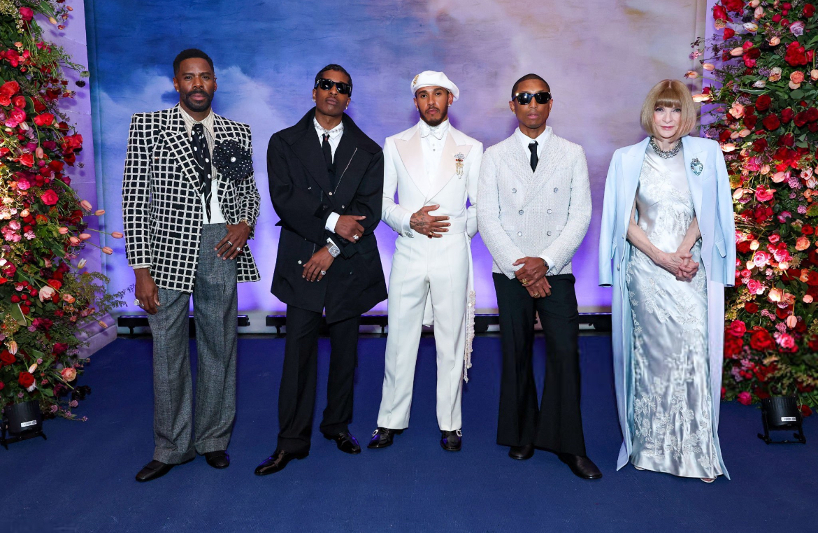 Colman Domingo, A$AP Rocky, Lewis Hamilton, Pharrell Williams e Anna Wintour posam juntos no tapete azul do Met Gala 2025. Os quatro co-anfitriões masculinos usam trajes elegantes que refletem o tema “Superfine: Tailoring Black Style”: Colman está com um blazer quadriculado em preto e branco com uma flor grande na lapela; A$AP Rocky veste um sobretudo preto estilizado com camisa branca e gravata; Lewis Hamilton está de branco dos pés à cabeça, com um terno de alfaiataria refinada e boina combinando; Pharrell usa um casaco branco com calça preta e óculos escuros. Anna Wintour, ao lado deles, veste um longo vestido prata claro com estampas florais e capa azul clara, mantendo seu clássico corte chanel.