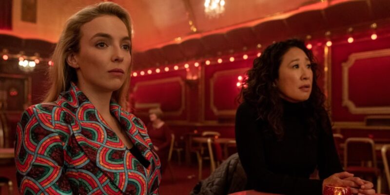 Cena de Killing Eve. Na imagem, duas moças jovens estão sentadas lado a lado em um salão de paredes vermelhas. À esquerda, está a personagem Villanelle, uma mulher branca que usa um terno com estampa rosa e azul. Com cabelos loiros, ela não usa acessórios e olha serenamente para o horizonte. À direita, está Eve Polastri, mulher de ascendência sul-coreana e cabelos escuros cacheados, que veste uma blusa preta de gola alta e mangas cumpridas. A personagem também olha para o horizonte, mas com feição preocupada.]