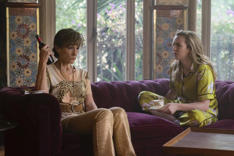 Cena de Killing Eve. Uma moça branca de cabelos loiros e uma senhora branca sentam em um sofá roxo. Atrás delas, há paredes com estampa florida e janelas. A senhora, com olhar desolado, encara o chão enquanto segura uma garrafa de vinho. Com cabelos grisalhos e presos em um coque, ela veste uma blusa com a imagem de uma onça desenhada e um cinto sobre ela. A calça é nude e possui pequenos pontos dourados. A moça loira conversa com a senhora, enquanto segura uma xícara de chá apoiada em um pires. Ela veste um pijama amarelo-limão com flores brancas estampadas. O dia aparenta estar ensolarado.]