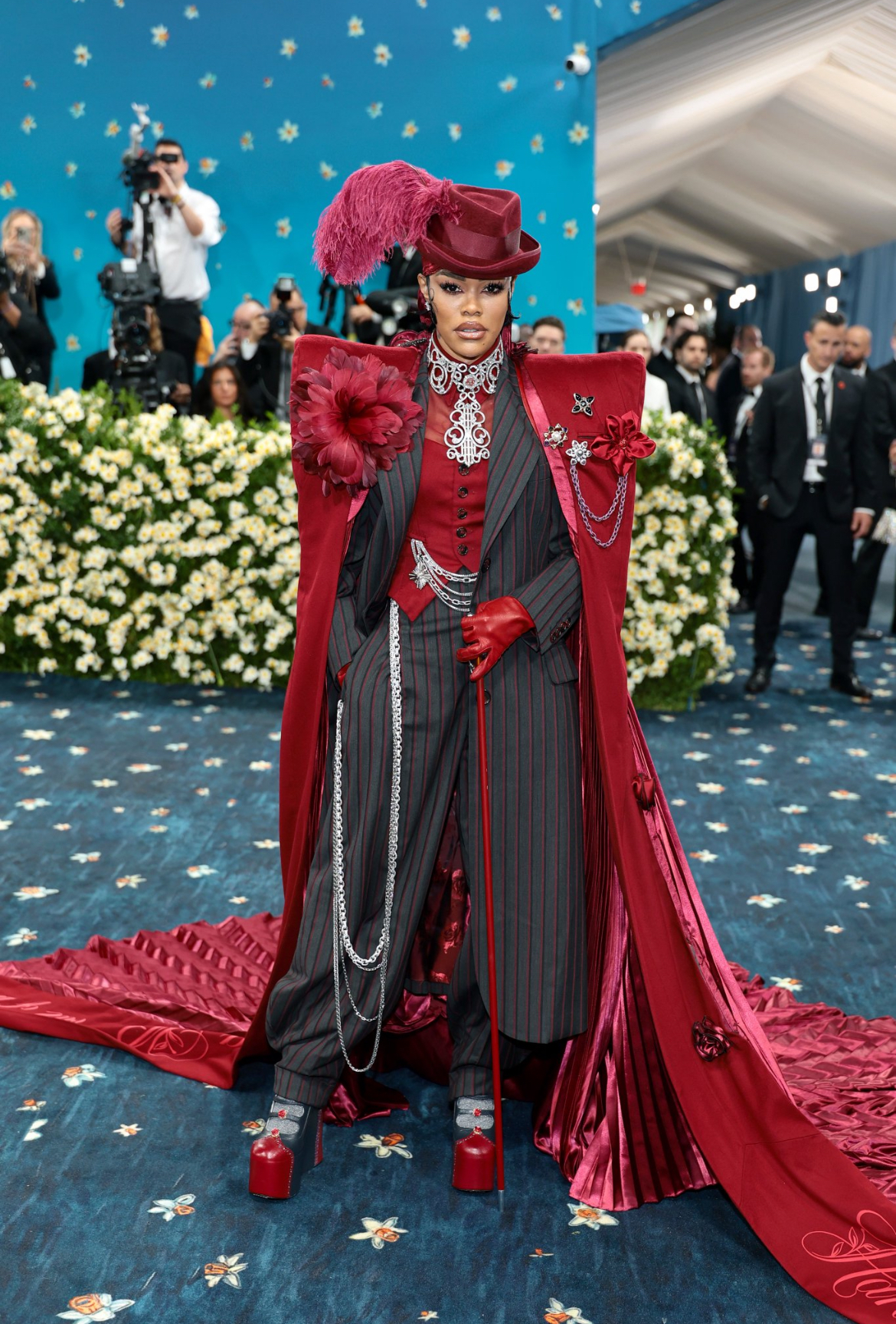 A cantora e atriz Teyana Taylor posa no tapete azul florido do Met Gala 2025 com um look ousado, teatral e rico em referências históricas. Ela usa um conjunto de alfaiataria com listras verticais em preto e vinho, combinado a um colete vermelho, luvas de couro, gravata rendada e sapatos plataforma vermelhos. O visual é finalizado com uma longa capa de cetim vermelho escuro plissado, chapéu com pluma, broches, correntes e uma flor volumosa presa ao ombro. Ao fundo, fotógrafos e convidados observam enquanto Teyana domina a cena com atitude e presença marcante.