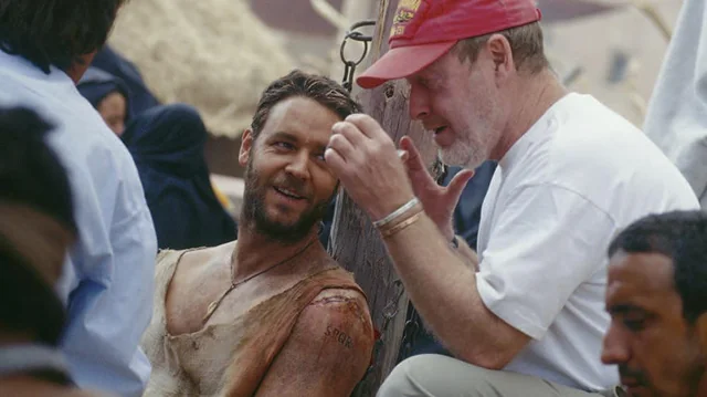 Cena de bastidores do filme GladiadorNa imagem, Russell Crowe, caracterizado como Maximus com roupas sujas e ferimentos cenográficos no ombro, conversa e sorri com o diretor Ridley Scott nos bastidores do filme. Scott, um homem mais velho de barba branca e boné vermelho, gesticula com as mãos enquanto fala. A cena se passa em um set ao ar livre, com figurantes ao fundo.