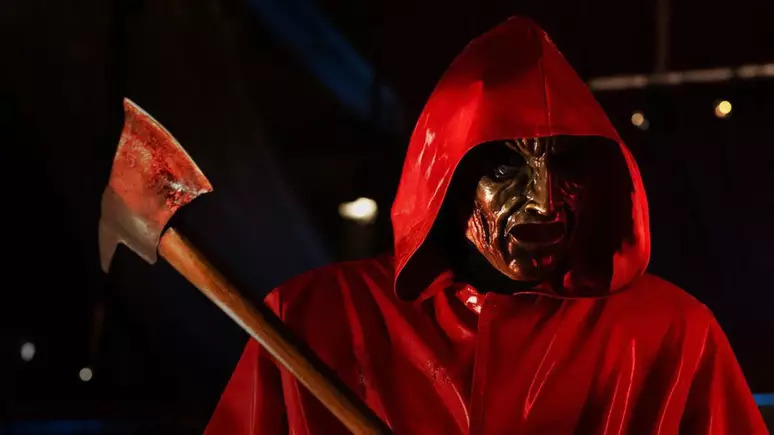 Cena do filme Rua do Medo: Rainha do Baile Na imagem, o assassino do filme encara alguém para matar. Ele segura um machado com cabo de madeira, que está sujo de sangue na lâmina. O personagem usa uma capa de chuva vermelha e uma máscara dourada semelhante à do teatro grego. 
