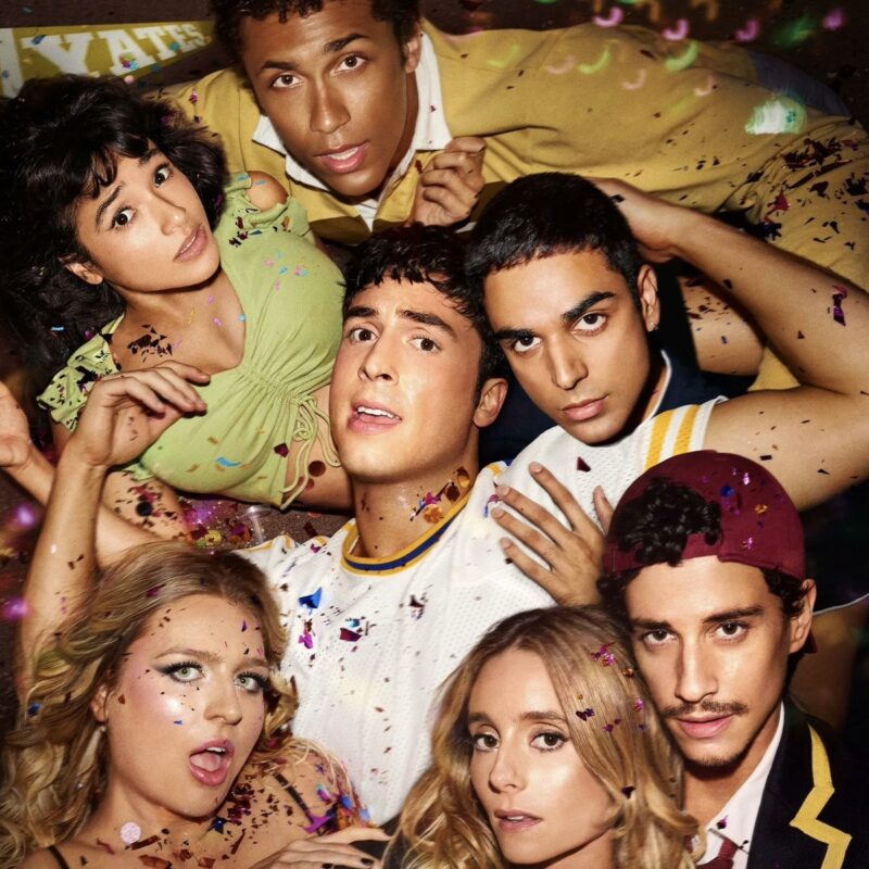 Imagem promocional da série Overcompensating. A foto mostra um close-up de um grupo de seis jovens adultos, em um ambiente colorido. A perspectiva é ligeiramente distorcida, como se fosse tirada de baixo para cima, e a cena é vibrante e caótica, com confetes coloridos espalhados sobre eles e no ar. No centro, Benny, um jovem de camiseta esportiva branca olha para cima com uma expressão de surpresa. Ao redor dele, os outros cinco o encaram ou olham para a câmera com expressões variadas: à esquerda, Carmen, uma jovem de cabelo escuro e blusa verde tem um olhar preocupado; acima, Gabe, um jovem de jaqueta amarela olha intensamente para a câmera; à direita de Benny, Miles, um jovem também encara a câmera com seriedade. Na parte inferior da imagem, há Hailee, uma jovem loira com a boca aberta, e outra jovem loira, Grace, com um olhar sério e direto, e um jovem, Peter, com bigode fino e um boné vermelho virado para trás. A iluminação é quente e difusa, com luzes desfocadas ao fundo, reforçando a atmosfera de festa.